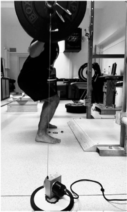 A barbell on a squat rack with a linear position transducer attached — the cable connects to the bar and feeds real-time velocity data to a nearby device